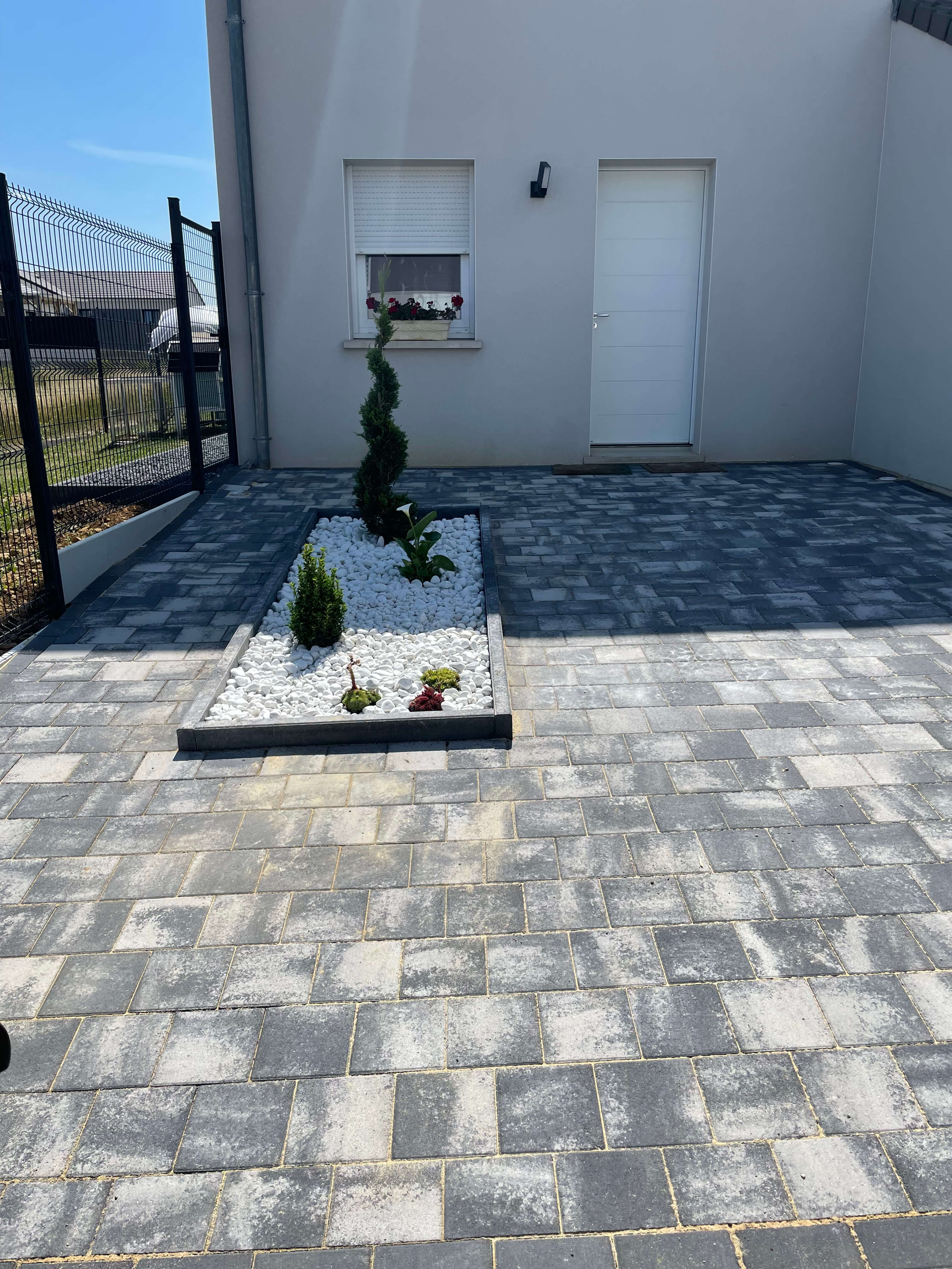Entrée de garage en pavé 20x20 Kronimus colorie noir blanc nuancé et décoration faite en bordure avec cailloux déco blanc et arbuste.